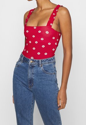 Vrouw met een rood mouwloos topje met witte madeliefjesprints en gerimpelde bandjes, gecombineerd met een blauw spijkerbroek met hoge taille.