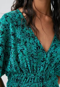 Robe vert canard avec un décolleté en V, dotée d'un tissu texturé et de taches noires. La taille est élastiquée pour un ajustement près du corps.