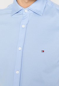 Camisa de algodón azul claro con botones, tejido texturizado, dos botones blancos visibles y un pequeño logo bordado en rojo, blanco y azul.