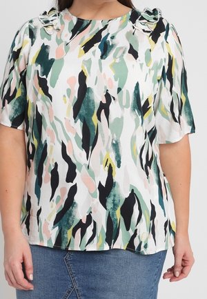 Vrouw die een blouse met korte mouwen en gerimpelde schouders draagt, met een groen, zwart, roze en geel abstract patroon en een blauwe spijkerrok.