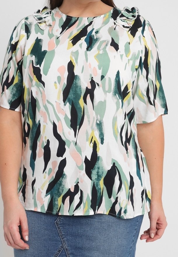 Femme portant un chemisier à manches courtes avec des épaules volantées, présentant un motif abstrait vert, noir, rose et jaune et une jupe en jean bleue.