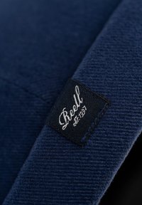 Tela de color navy con un tejido texturizado presenta una etiqueta negra cosida con texto en cursiva blanca, que detalla la marca y el año de establecimiento.
