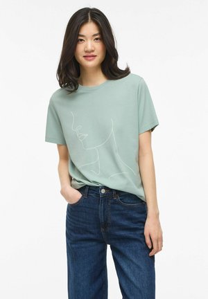 Jeune femme aux cheveux foncés portant un T-shirt vert clair avec un dessin au trait blanc représentant un visage, associé à un jean bleu foncé.