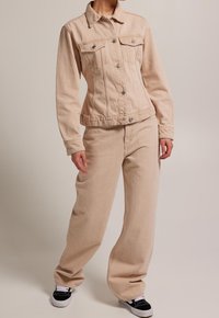 Beige denimjacka med två bröstfickor, knappknäppning framtill och insvängd midja. Kombinerad med löst sittande matchande beige byxor och svarta sneakers.