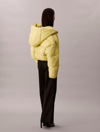 Κοπής κίτρινο μπουφάν με κουκούλα και ραφές, σε συνδυασμό με μαύρα παντελόνια και μποτάκια με τακούνι, ορατό από πίσω.