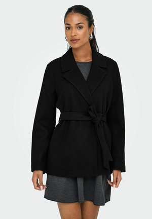 Abrigo negro con cinturón, cuello, mangas largas y una textura suave. Llevado sobre un vestido gris, resaltando su diseño entallado.