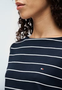 Μπλε ναυτικό πουκάμισο με άσπρες οριζόντιες ρίγες, με μικρή λεπτομέρεια λογότυπου σε κόκκινο, άσπρο και μπλε στο στήθος. Μαλακό, λεία υφάσμα.