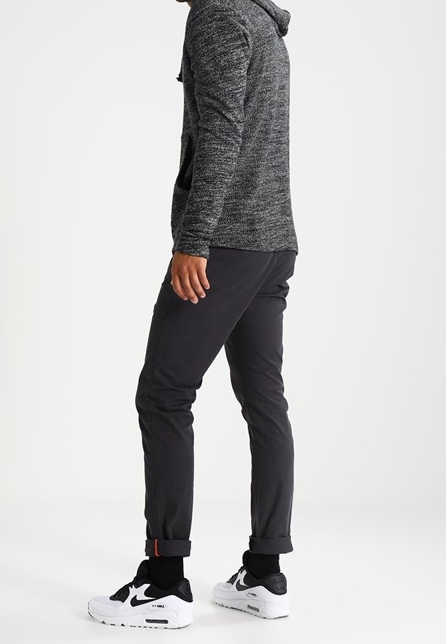 Homme portant un sweat à capuche gris, un pantalon noir avec revers, des chaussettes noires et des baskets blanches Nike Air Max, debout sur un sol blanc.