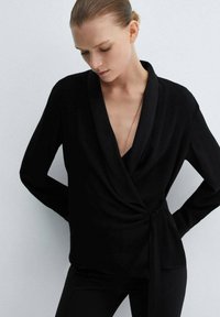 Top nero a maniche lunghe con scollatura a V profonda. Realizzato in tessuto liscio e leggero con dettaglio annodato sul lato.