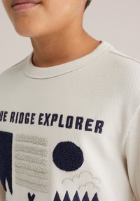 Světle béžová mikina má texturovanou tkaninu, kulatý výstřih a tmavě modré vyšívané akcenty s geometrickými vzory a nápisy.