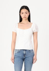 Blouse en coton blanche avec des manches courtes bouffantes, un décolleté carré et des broderies florales. Présente un détail de boutons sur le devant. Associée à un jean bleu.
