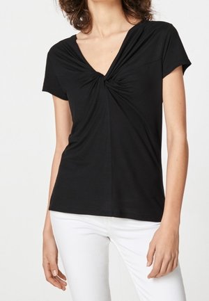 Femme portant un haut noir à manches courtes avec un col en V et un détail froncé au niveau de la poitrine, associé à un pantalon blanc.