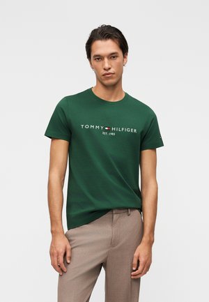 Νέος άνδρας που φοράει σκούρο πράσινο T-shirt Tommy Hilfiger με λογότυπο και μπεζ παντελόνι, στέκεται μπροστά σε απλό ανοιχτό φόντο.