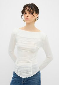 Vit långärmad topp med fyrkantig halsringning, veckad front och slät, lätt textur, tillsammans med blå denimjeans.