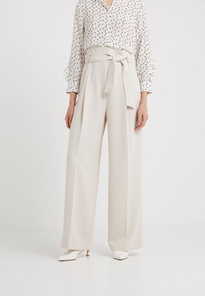 Kvinde iført cremede, højtaljede, vide bukser med taljebælte, kombineret med en hvid bluse med små sorte print og hvide hæle.