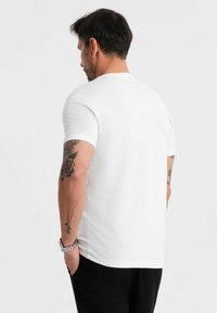 Camiseta blanca de manga corta en un ajuste clásico, hecha de tejido suave. Presenta un cuello redondo y tatuajes visibles en el brazo derecho.