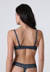 Donna che indossa un reggiseno in pizzo blu scuro e un perizoma abbinato, mostrata di spalle su uno sfondo grigio chiaro uniforme.