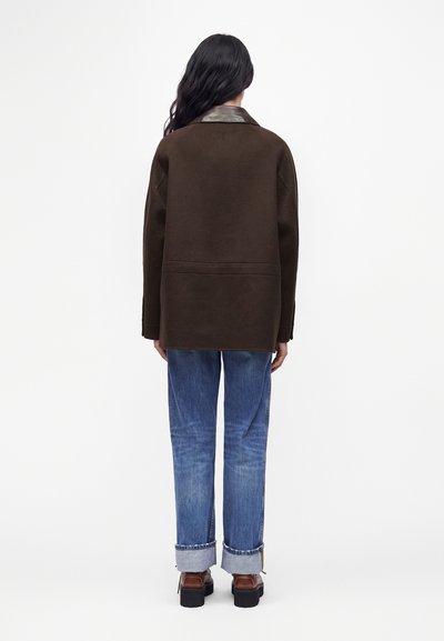 Femme aux longs cheveux foncés portant une veste marron foncé avec un col en cuir, un jean bleu roulé et des chaussures marron, debout de dos.