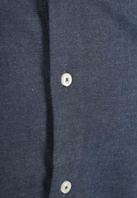 Donkerblauw hemd met zichtbare structuur. Bevat een rij knopen in een lichte kleur met een subtiele glans en een smalle knoopsluiting.