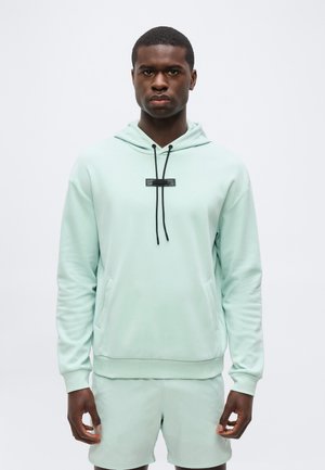 Mann med lys mintgrønn hettegenser med svarte snorer og matchende shorts, står foran en ensfarget hvit bakgrunn.