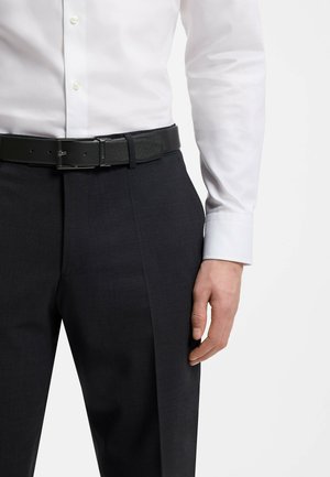 Mann trägt ein weißes Hemd, in schwarze formelle Hosen mit einem schwarzen Ledergürtel gesteckt, Hand seitlich am Körper.