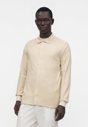 Uomo che indossa una camicia trapuntata beige a maniche lunghe con bottoni e pantaloni bianchi, in piedi su uno sfondo bianco semplice.