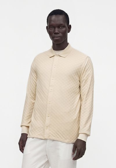 Homme portant une chemise matelassée beige à manches longues boutonnée et un pantalon blanc, debout devant un fond blanc uni.