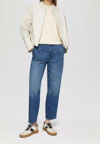 Jasnobeżowa kurtka bomberka, kremowy top, niebieskie jeansy z wysokim stanem oraz białe trampki z czarnymi wstawkami i brązowymi podeszwami. Gładki i casualowy wygląd.