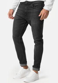 Jeans de mezclilla negra con un corte slim, que cuentan con un diseño clásico de cinco bolsillos y puños enrollados, combinados con zapatillas blancas.
