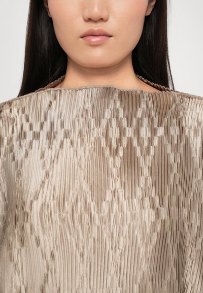 Textured, pleated fabric top in light beige with a round neckline and intricate, alternating patterns. Smooth and glossy finish.