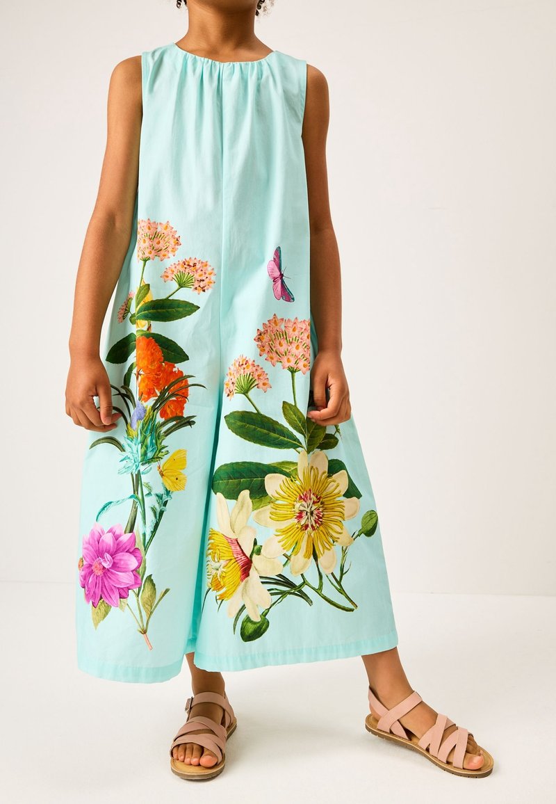 Enfant portant une robe sans manches bleu clair avec de grands imprimés floraux et papillons colorés, et des sandales roses sur un fond clair.