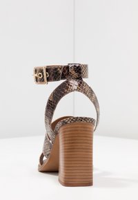 Öppen tå-högklackade sandaler med ett ormskinnsmönster, en tan-färgad konstläderrem, ankelspänne och en rektangulär, räfflad brun klack.