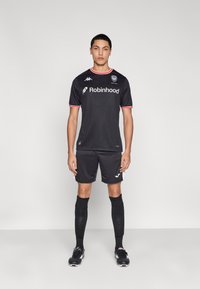 Equipación de fútbol negra con cuello redondo y detalles en rojo, mangas cortas y el logo "Robinhood" en el frente. Pantalones cortos y calcetas negras completan el conjunto.