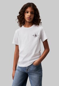 White cotton t-shirt with short sleeves, featuring a small printed logo "Calvin Klein Jeans" in grey. Paired with light blue denim jeans.