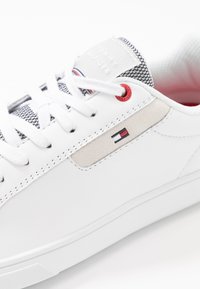 Baskets blanches avec des lacets blancs, détail œillet rouge, patch logo Tommy Hilfiger cousu et languette texturée avec texte de la marque.