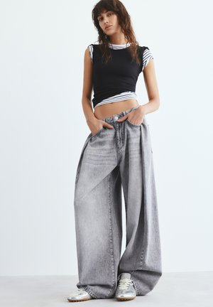 Fiatal nő áll, kezét a zsebében tartja, túlméretezett szürke farmernadrágot, fekete ujjatlan felsőt csíkos ing fölött, valamint ezüst tornacipőt visel.