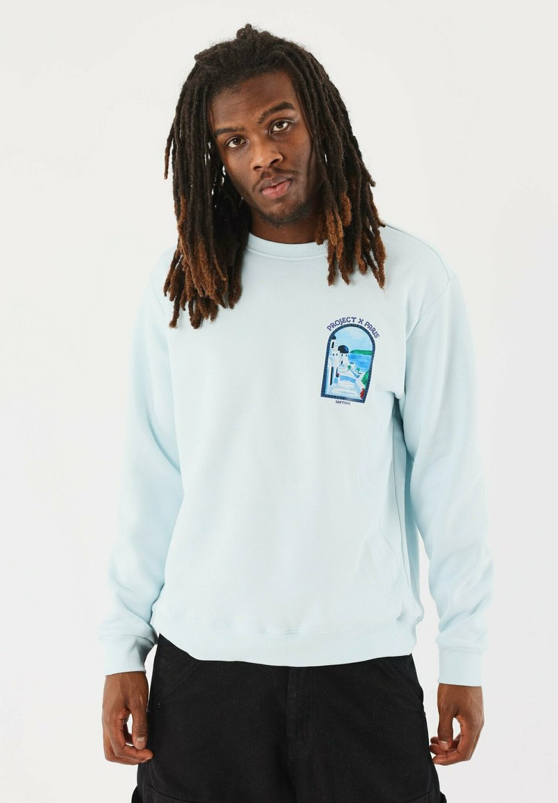 Sudadera azul claro de material suave, con un pequeño gráfico bordado en el lado izquierdo del pecho que incluye texto y detalles del diseño.