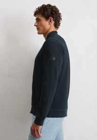 Dark green lightweight zip-up jacket with ribbed texture, side pockets, and a black logo patch on the left sleeve. Paired with light blue jeans.