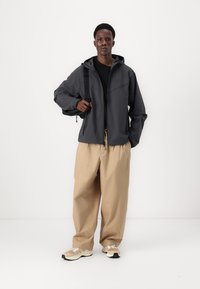 Donkergrijze waterdichte jas met een rits aan de voorkant, een aangehechte capuchon en elastische manchetten, gecombineerd met losse beige broek en lichte sneakers.