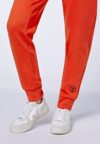 Oranger Fleece-Sweatpants mit gerippten Bündchen, featuring ein kleines Logo am linken Knöchel. Kombiniert mit weißen und grauen Sneakers.
