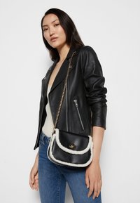 Svart läder crossbody-väska med vit konstpälskant, gyllene detaljer och kedja, som har en rundad klaffdesign.