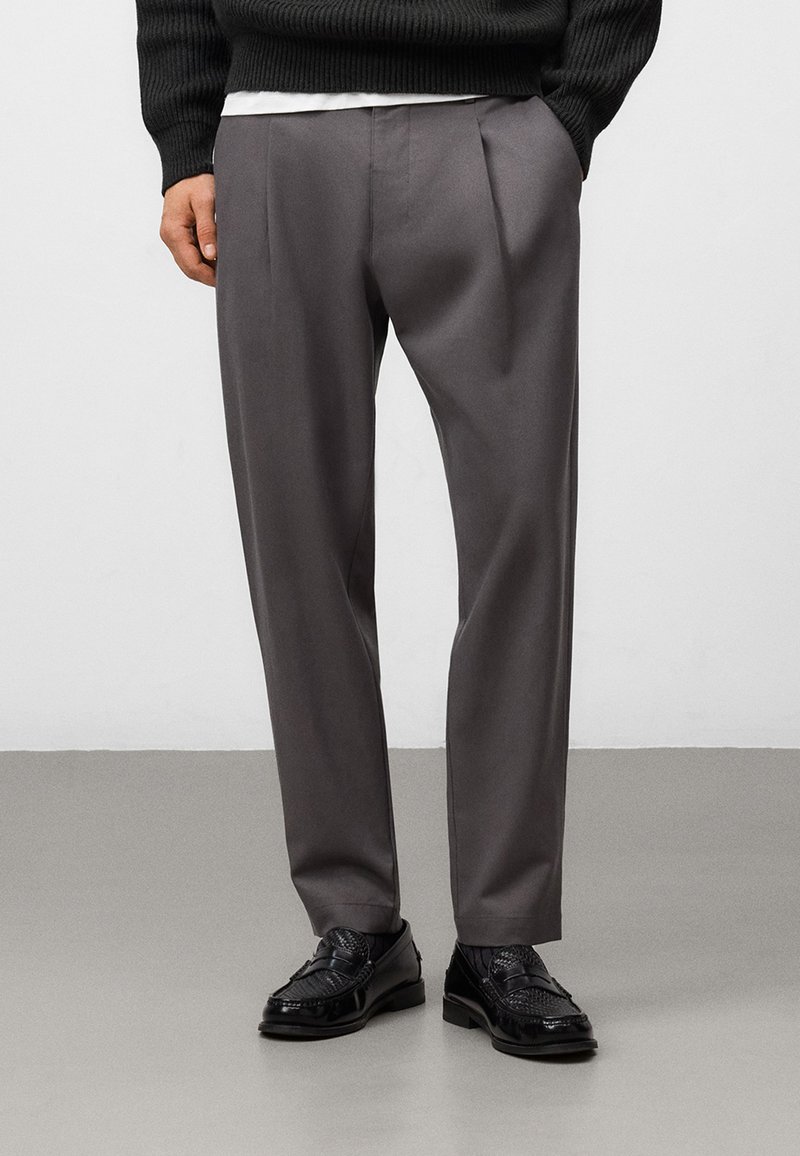 Grå veckade byxor tillverkade av mjukt tyg, med en skräddarsydd passform, bärs med svarta texturerade loafers och en mörk ribbad tröja.