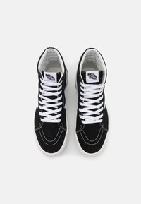 Czarne zamszowe trampki typu high-top z białymi sznurowadłami, kontrastowym przeszyciem i białą gumową podeszwą. Na języku znajduje się logo Vans.