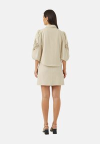 Beige jakke med puffede ærmer, broderede detaljer på ærmerne, sammen med en matchende tætsiddende nederdel og sandaler med blokhæle.
