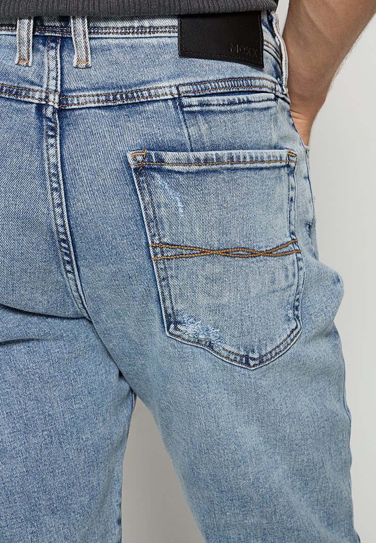 Hellblaues Denim-Jeans mit sichtbarem Hintertaschen und braunen Nähten, getragen von einer Person, deren Hand in der Nähe der Taille ruht.