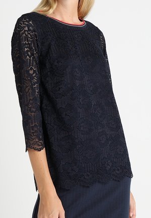 Marineblauwe blouse van bloemenkant met driekwart mouwen en een geschulpte zoom, gedragen met een donkere gestreepte rok.