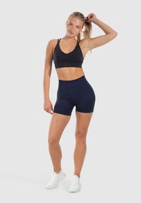 Černá sportovní podprsenka s výstřihem do V a nařaseným středem, spárovaná s navy šortkami s vysokým pasem. Modelka má na sobě bílé sportovní boty.