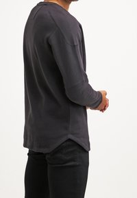 Camisa de manga larga gris oscuro con dobladillo redondeado, tejido texturizado y detalles en los hombros; combinada con pantalones negros. Vista trasera.