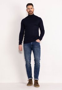 Marineblauwe coltrui, slim-fit spijkerbroek, casual bruine veterschoenen, model staand tegen een effen achtergrond.