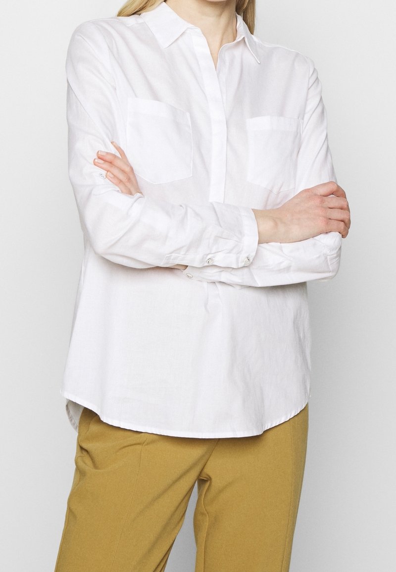 Personne portant une chemise blanche à manches longues boutonnée avec des poches poitrine et un pantalon couleur moutarde, les bras croisés sur le torse.